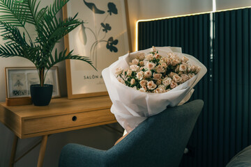 Bouquet of delicate pink roses wrapped in white paper, placed on a chair in a cozy modern interior with wooden desk and framed art