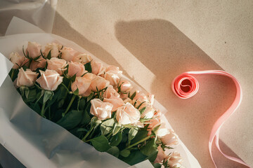 Elegant bouquet of pale pink roses wrapped in white paper with a pink ribbon roll nearby on light textured surface in sunlight
