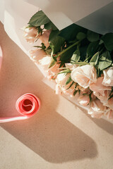 Elegant bouquet of pale pink roses wrapped in white paper with a pink ribbon roll nearby on light textured surface in sunlight