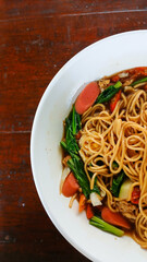 Top view of a white bowl filled with stir-fried noodles, sausage slices, leafy greens, cabbage, and chili in a savory sauce, placed on a rustic wooden table.