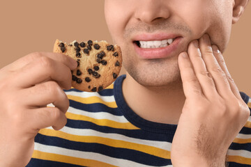 Young man with cookie suffering from toothache on brown background, closeup