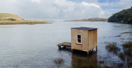 A small, wooden shack sits on stilts in the Awakino river, connected to a dock. Used for...