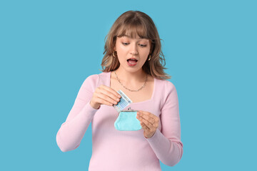 Surprised young woman putting money in purse on blue background