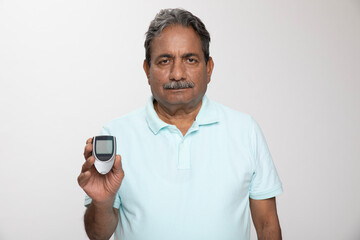 Blood sugar machine. Portraits of indian senior man showing blood glucose meter while standing grey...