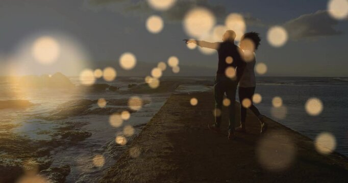 Couple walking stone pier reaching midpoint causing golden bokeh lights pointing horizon for travel