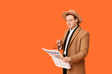 Young man in hat, coat and with newspaper on orange background