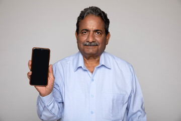 Portrait of indian happy senior man showing mobile phone screen while isolated on grey background Indian Cheerful senior man earing formal shirt and using by phone