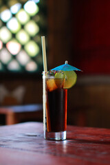 Classic Cuba Libre served in a tall straight glass with lime on the rim and ice cubes. Set on rustic wooden table with natural light. No people, refreshing rum cocktail.