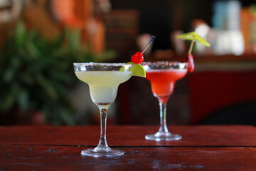 Two margaritas on rustic wooden table: vibrant strawberry with paper umbrella, classic with lime wedge and cherry garnish. Natural light, no people, refreshing cocktails.