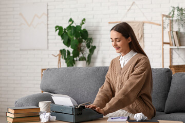 Beautiful happy female writer with typewriter, books and notebook sitting on sofa at home