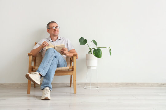 Old man reading book in armchair near white wall