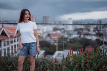 Asian woman standing in a rooftop garden, holding a tablet and gazing at a stunning cityscape at dusk, dressed in a casual white t-shirt and denim shorts