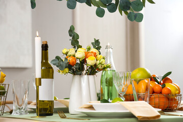 Festive table setting with citrus fruits, bottles of drinks, flowers and burning candles in room. Closeup