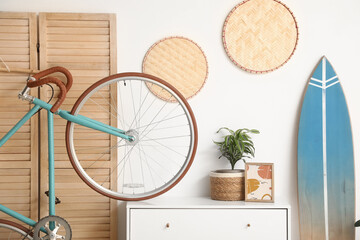 Naklejka na ściany i meble Interior of living room with chest of drawers, bicycle and surfboard near white wall