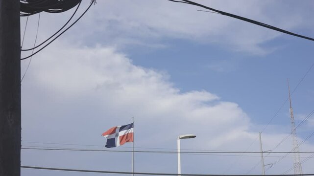 dominican republic flag waving under a cloudy blue sky in an urban setting, with visible street lights and power lines crossing the frame, canon eos R c-log color 8bits