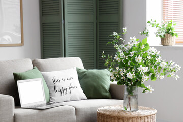 Sofa, table, modern laptop and vase with blooming jasmine flowers in living room, closeup