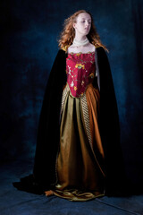 A red-haired Renaissance woman wearing an embroidered silk ensemble against a studio backdrop