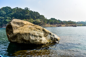 Large boulder resting in clear Dawki river water, surrounded by lush trees.