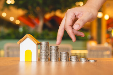 A wooden house model with rising coins, guided by an investor's finger, symbolizes investment. It...