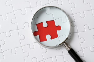 Close-up View of a Magnifying Glass Focused on a Single Red Puzzle Piece Amidst White Puzzle Pieces on a Light Background
