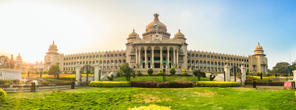 Panoramic view of Vidhana Soudha, the iconic government building in Bengaluru, India, captured during golden hour. The lush gardens and clear sky frame the historic architecture beautifully