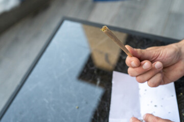 Man holds a cannabis joint above a dark marble table, with rolling papers and crushed herb nearby. The mood is casual and calm, highlighting recreational use.