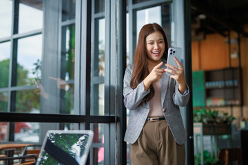 Fototapeta premium Asian businesswoman engaging in a video call on her smartphone, standing by the window in a cozy cafe, radiating confidence and professionalism
