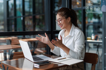 Fototapeta premium Young Asian businesswoman smiling and gesturing while engaging in a video call on her laptop at a cozy cafe, embodying modern remote work