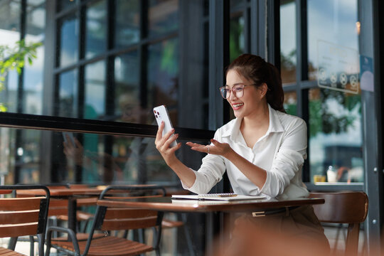 Young cheerful Asian businesswoman having video call using smartphone while working remotely in outdoor cafe