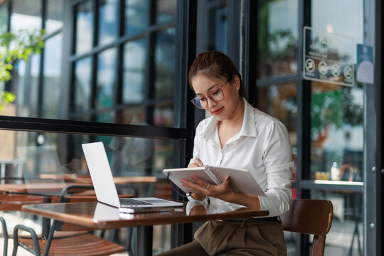 Asian businesswoman wearing eyeglasses taking notes in notebook while working on laptop in cafe