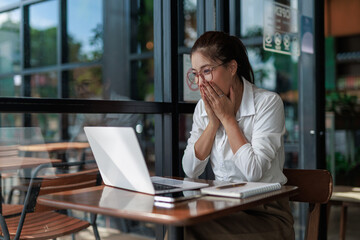 Fototapeta premium Asian businesswoman covering her mouth with her hands while looking at her laptop in a cafe, expressing surprise and shock