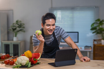 Young man wearing an apron, holding a green apple and using a tablet in a kitchen with fresh fruit and vegetables on the table
