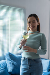 Happy asian woman enjoying glass of chilled white wine standing in living room with window blinds in background