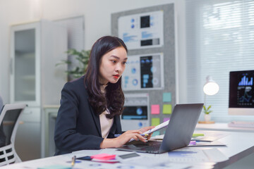 Asian businesswoman working diligently, comparing information from documents and a laptop in a sleek, modern office environment