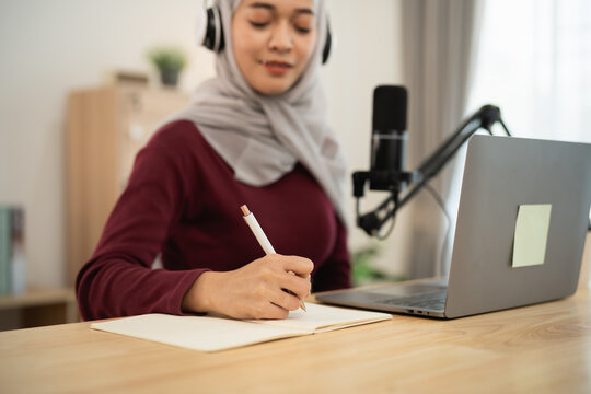Young woman in casual attire wearing headphones while writing notes in cozy workspace with laptop and microphone for podcast or online content creation - Powered by Adobe