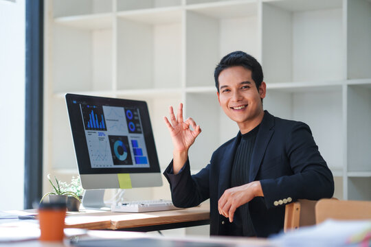 Happy businessman showing ok sign while analyzing positive financial charts on computer at office desk