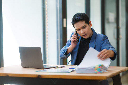 Young entrepreneur working with laptop and documents at office desk while talking on mobile phone