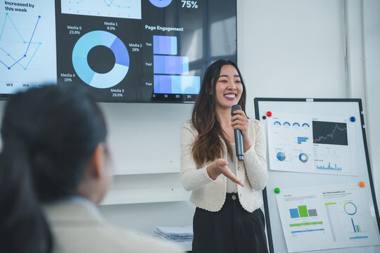 Young Asian businesswoman leading a presentation using financial charts on screen and flip chart, explaining diagrams and smiling