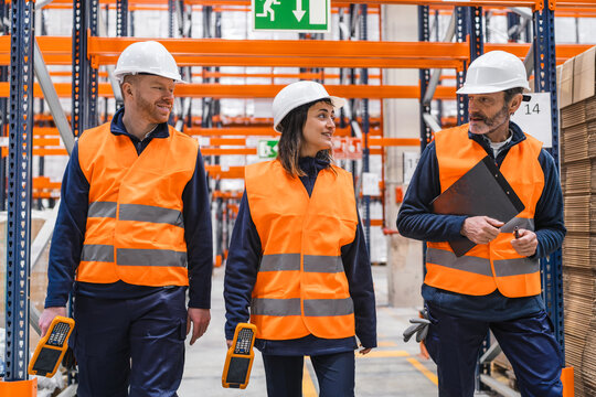 Warehouse workers wearing safety vests and helmets are walking through the facility, discussing logistics and using barcode readers