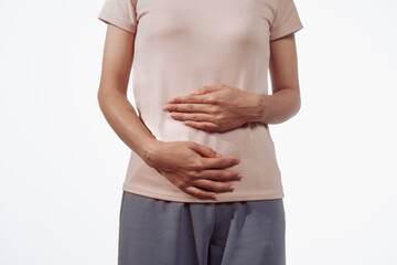 A confident young Asian woman in a light brown shirt stands against a white background, experiencing abdominal pain. Causes may include menstrual cramps, ovarian cysts, endometriosis