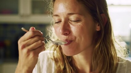 Smiling Caucasian woman with curly hair enjoys yogurt for breakfast in a bright kitchen. She holds a spoon and looks happy while eating. - Powered by Adobe