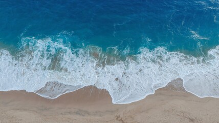 An aerial view looks down on a sandy beach where turquoise ocean waves are crashing and receding, leaving foamy white patterns on the sand. 