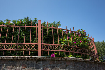 a stone fence with a forged or cast metal grating of reddish-brown color installed on top. A lush...