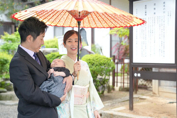 In the spring month of May, a one-month-old Japanese baby is held by his father in a suit. Beside them, the baby's mother, a woman in her twenties wearing a kimono, smiles while holding an umbrella.