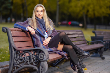 Cute Tranquil Winsome Caucasian Beautiful Blond Young Woman In Long Striped Coat Posing in Autumn Park On Bench Outdoors