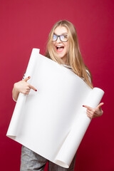 Cute Winsome Positive Caucasian Young Blond Girl In Glasses Posing With White Advertising Copy Space Rolling Paper List Over Trendy Burgundy Background While Pointing With Finger.