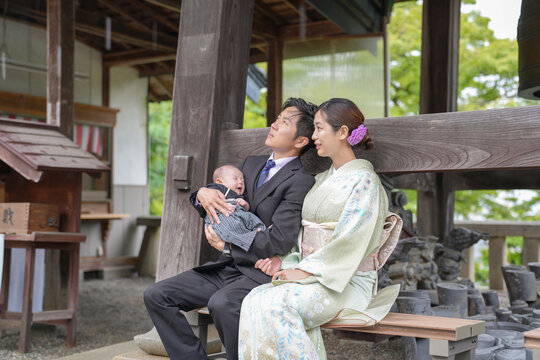 On a spring day in May at a Japanese temple, a one-month-old Japanese baby in hakama is held by a mother in kimono and a suited man, both in their twenties, seated on a bench, watching the rainy sky.