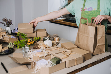 Person places kraft box into a paper bag at a workspace with handmade soap packaging. Sustainable...