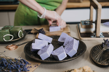 Person in green apron stamps a lavender soap bar by hand on a wooden board. Handmade soap branding, artisan process, small batch skincare, craft identity