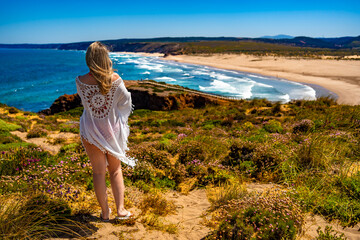 Beautiful middle aged woman standing in flowing dress at viewpoint on sandy beach Bordeira Praia in...